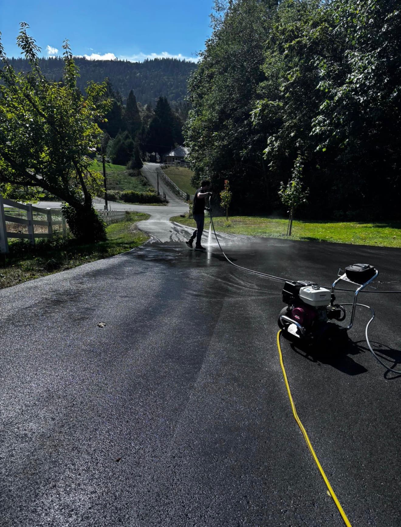 A Yectli worker pressure washing a paved driveway, with the washer and hose in the foreground.